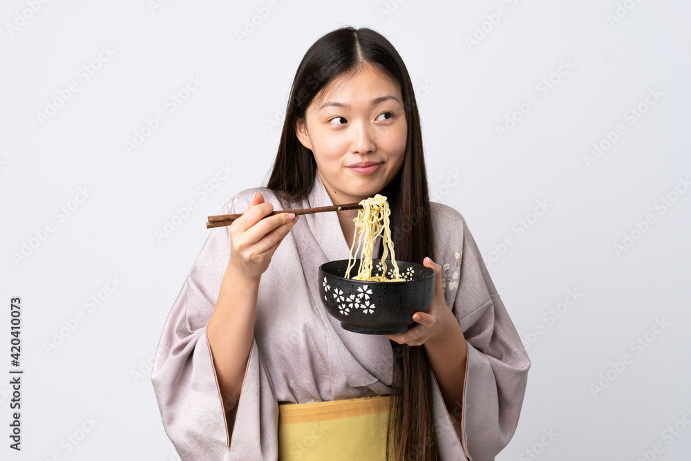 Young Chinese girl wearing kimono over isolated white background holding a bowl of noodles with chopsticks Young Chinese girl wearing kimono over isolated white background holding a bowl of noodles with chopsticks