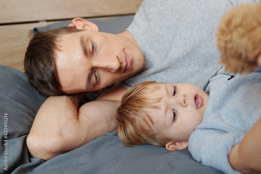 Young father lying on bed and resting together with his little son during sleep hour Young father lying on bed and resting together with his little son during sleep hour
