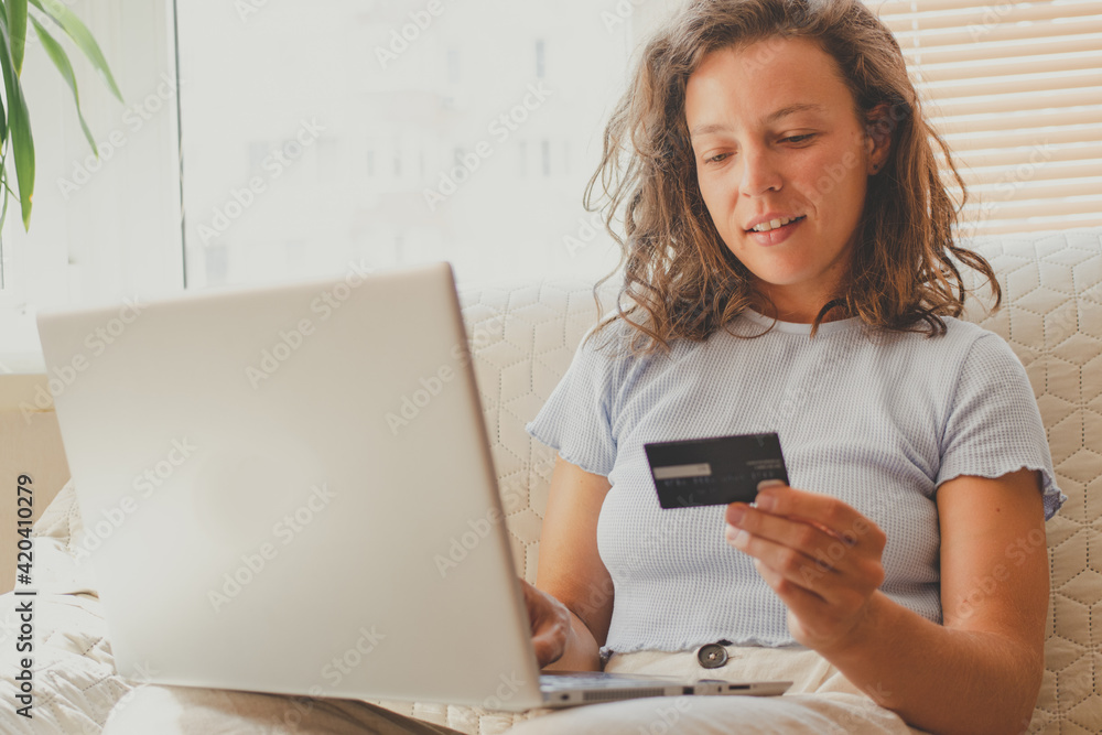 Young woman holding bank credit card, using laptop computer, entering card number, sitting on sofa at home. Making internet payment,learning payment system,choosing service, product in internet store Young woman holding bank credit card, using laptop computer, entering card number, sitting on sofa at home. Making internet payment,learning payment system,choosing service, product in internet store