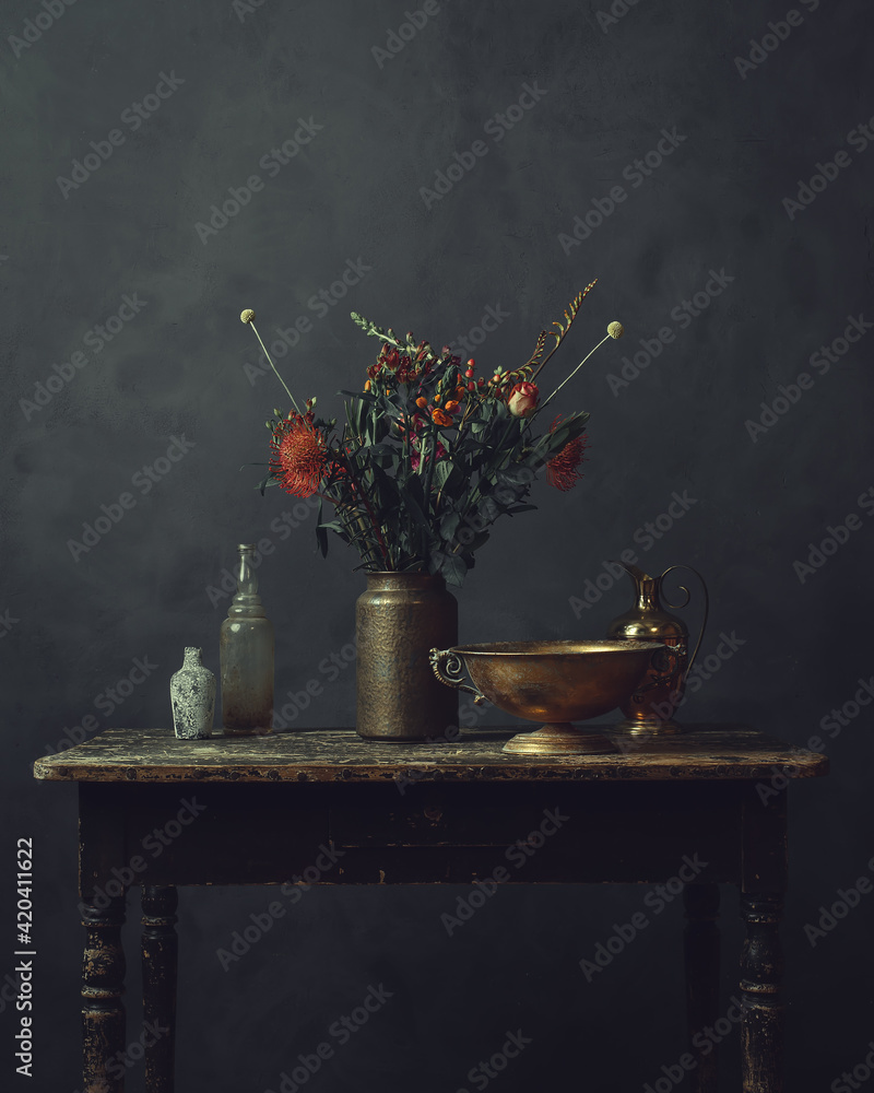 Old worn wooden table topped with a vase with flowers, a bronze bowl, pitcher and two glass bottles.