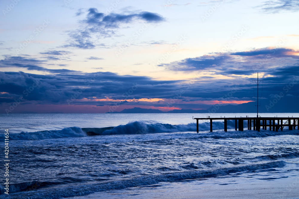 View from beach to water of sea, waves with white foam, pierce and sky with clouds in a nice evening.