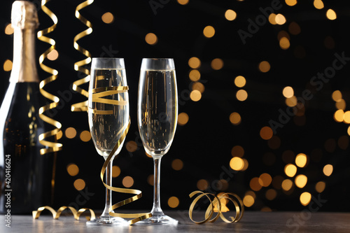 Glasses and bottle of champagne with serpentine streamers against blurred lights on black background. Space for text