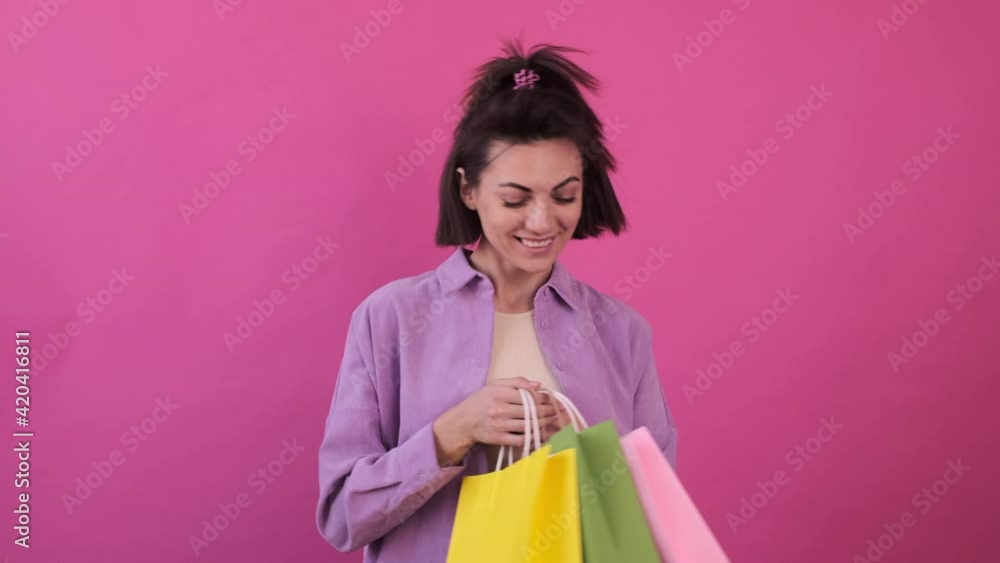 Caucasian woman in casual purple shirt on pink background happy dancing with colorful shopping bags
