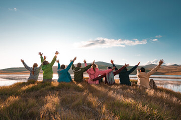 Big group of tourists sits with raised arms at sunset lake