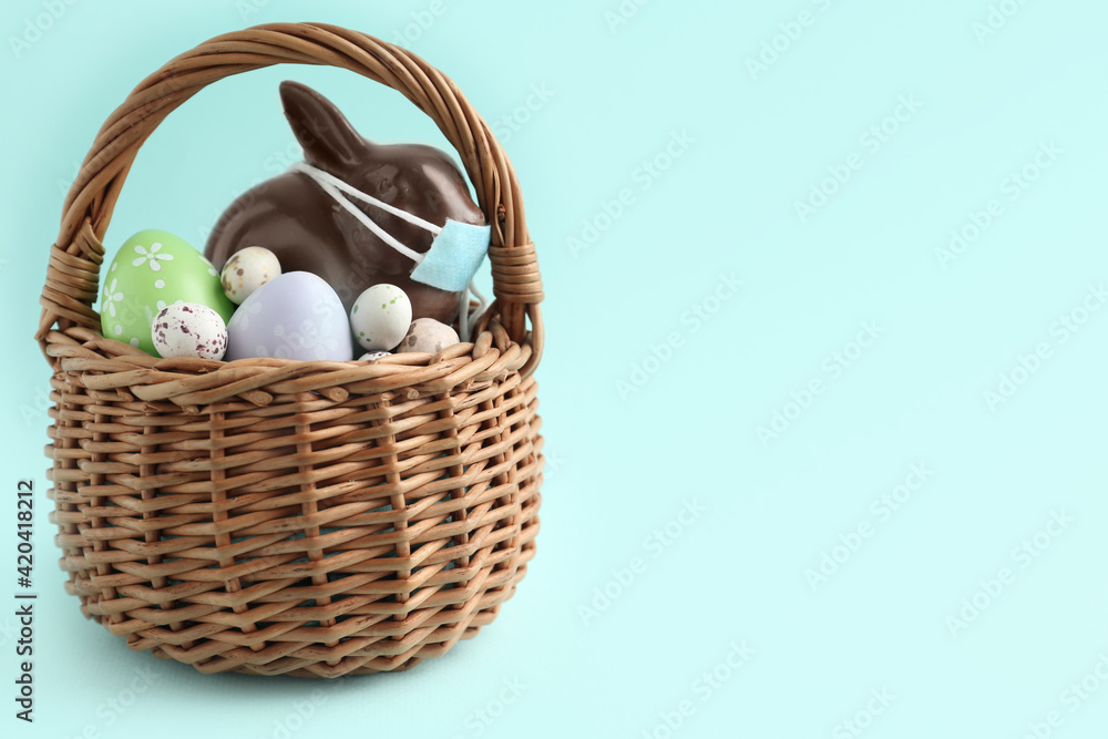 Chocolate bunny with protective mask and eggs in basket on light blue background, space for text. Easter holiday during COVID-19 quarantine