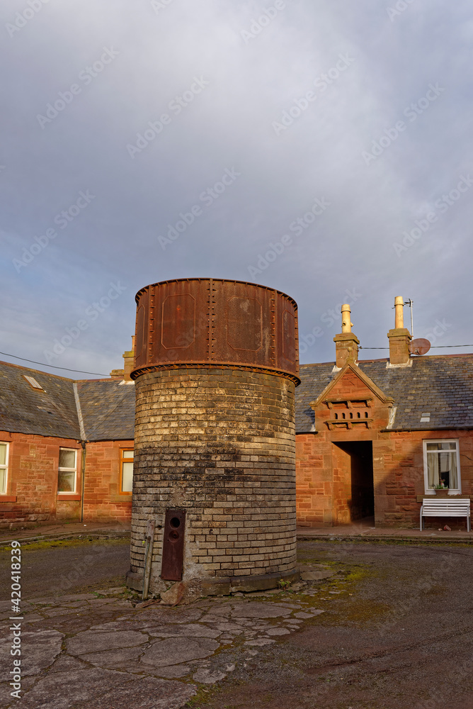 Fototapeta premium The Old Historical Iron Water Tower on a brick base in Fountain Square Auchmithie, with a small Doocot above the entrance of the central cottage,