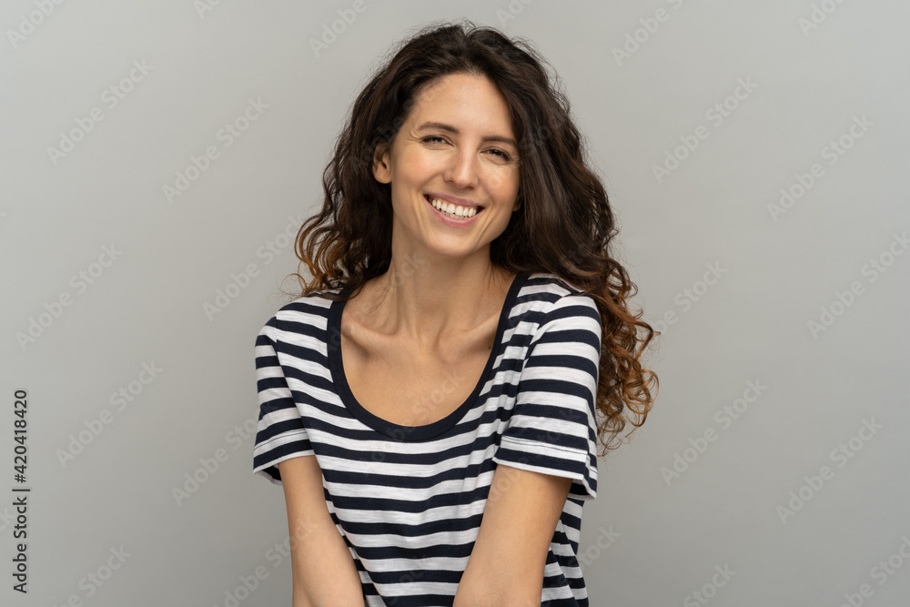 Fototapeta premium Portrait of European happy woman with curly hair and toothy smile, looking at camera. Pretty girl wear stripped t-shirt laughing, having fun standing isolated on studio grey background with copy space