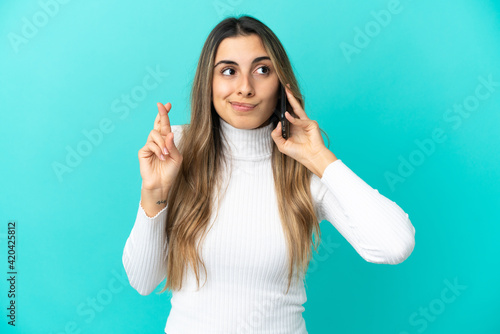 Young caucasian woman using mobile phone isolated on blue background with fingers crossing and wishing the best