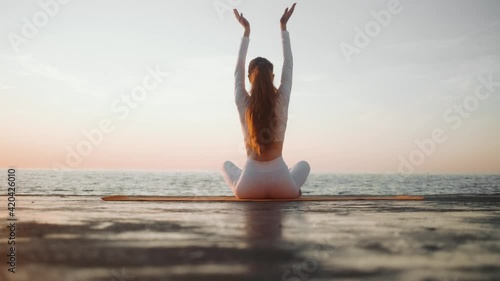 A back-view of a young redhead woman athlete wearing white sportsuit is doing yoga sitting outside near the sea in the morning