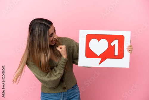 Young caucasian woman isolated on pink background holding a placard with Like icon and celebrating a victory