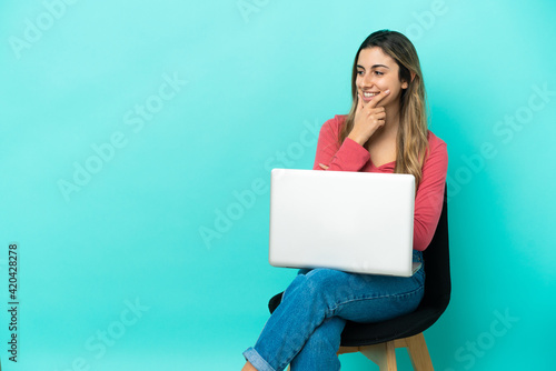 Young caucasian woman sitting on a chair with her pc isolated on blue background looking to the side © luismolinero