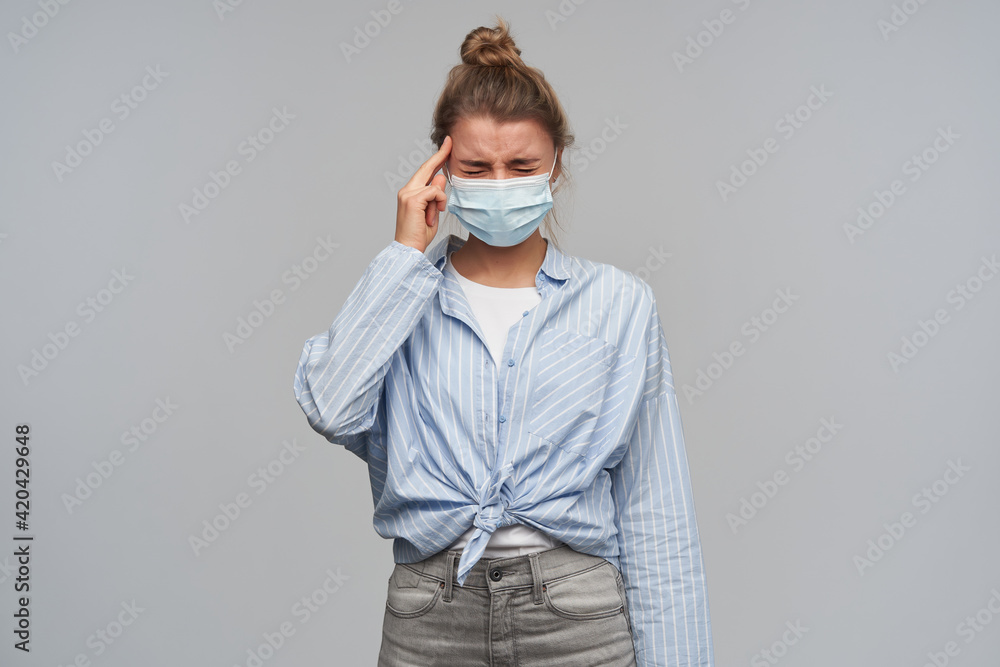 Stressed, unhappy woman with blond hair gathered in bun. Wearing striped knotted shirt and protective face mask. Touching her temple, have terrible headache. Stand isolated over grey background Stressed, unhappy woman with blond hair gathered in bun. Wearing striped knotted shirt and protective face mask. Touching her temple, have terrible headache. Stand isolated over grey background
