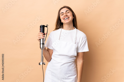 Woman using hand blender over isolated background laughing