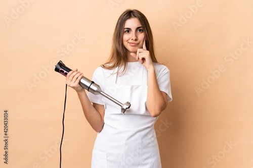 Woman using hand blender over isolated background and thinking