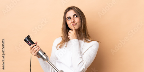 Woman using hand blender over isolated background having doubts while looking up