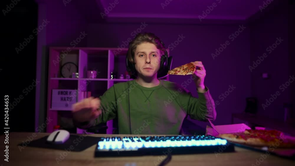 Hungry young male gamer plays online games at home at night and eats a delicious pizza out of a box, looks at the camera and uses a computer during dinner. Hungry young male gamer plays online games at home at night and eats a delicious pizza out of a box, looks at the camera and uses a computer during dinner.