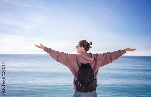 Young female traveler standing on the sea coast with back and arms raised