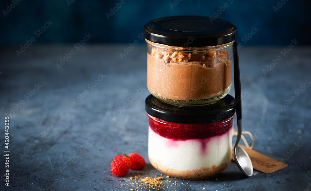 Two homemade easy cheesecakes in jars with chocolate mousse and raspberry curd sauce on a dark blue background. Healthy dessert. Selective focus. Homemade Raw Vegan Pudding. Two homemade easy cheesecakes in jars with chocolate mousse and raspberry curd sauce on a dark blue background. Healthy dessert. Selective focus. Homemade Raw Vegan Pudding.