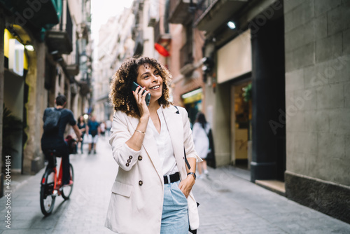 Pretty female tourist in stylish clothing walking at city street and discussing solo vacations experience during international smartphone conversation in roaming, happy girl using cell for talking © BullRun