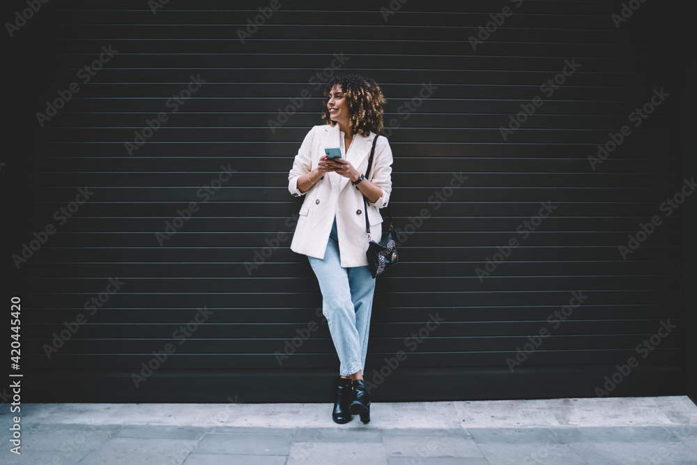 Trendy dressed female blogger with modern cellphone device in hand spending leisure at promotional urban setting, happy hipster girl with smartphone smiling near copy space area for fashion text