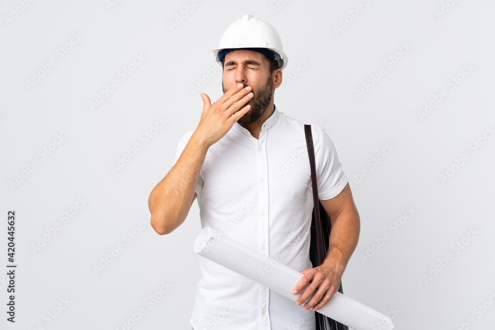 Young architect man with helmet and holding blueprints isolated on white background yawning and covering wide open mouth with hand Young architect man with helmet and holding blueprints isolated on white background yawning and covering wide open mouth with hand