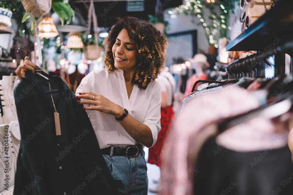 Smiling female standing near hangers with brand wear and laughing during Black Friday shopping with discounts, funny hipster girl enjoying pastime for update her wardrobe spending day in boutique - obrazy, fototapety, plakaty 