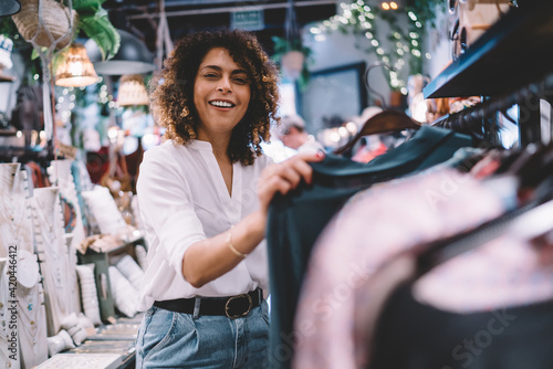 Portrait of joyful female customer enjoying weekend shopping in brand showroom, cheerful woman in trendy outfit buying stylish clothing and smiling at camera while posing near hangers with wear