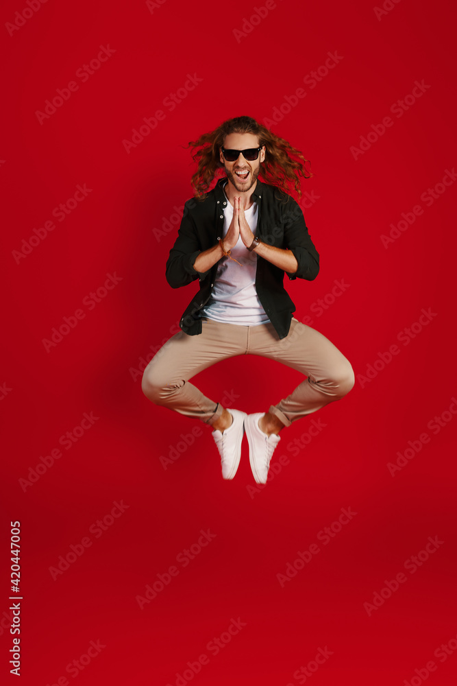 Full length of charming young man in casual clothing smiling and meditating while hovering against red background Full length of charming young man in casual clothing smiling and meditating while hovering against red background