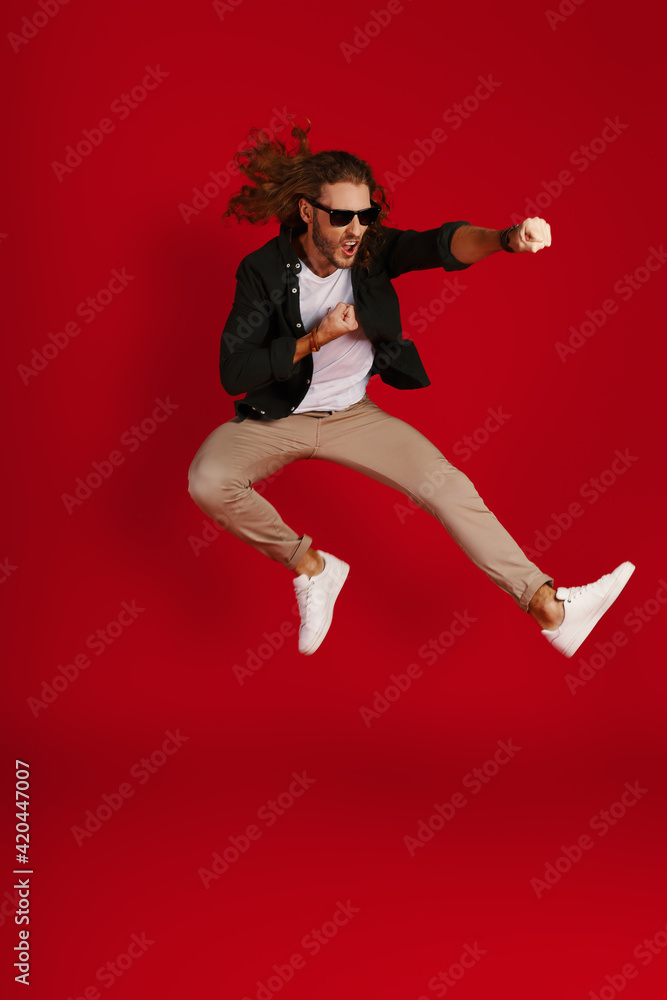 Full length of playful young man in casual clothing making a face and punching while hovering against red background Full length of playful young man in casual clothing making a face and punching while hovering against red background