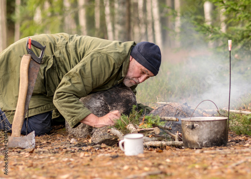 man in the forest trying to light a campfire, a tree branch with a pot over the campfire, blurred forest background, bonfire and smoke, autumn time