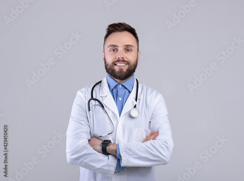 Portrait of positive male doctor posing with crossed arms and smiling at camera over grey studio background © Prostock-studio