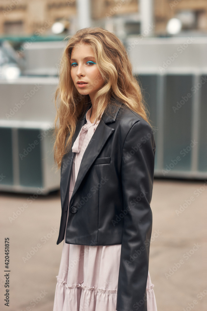 Fototapeta premium Young stylish teenager girl with blue eye arrows and long blonde wavy hair. Trendy fashionable female in pink dress, black leather jacket and high boots. Rock style woman outdoor portrait