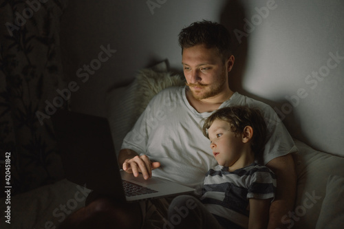 Father and son using laptop by night at home on the bedroom.