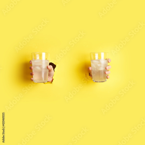 woman hands holding glasses with lemonade, lemon and ice in both hands through the hole in yellow paper. Summer lemonade concept. High quality photo