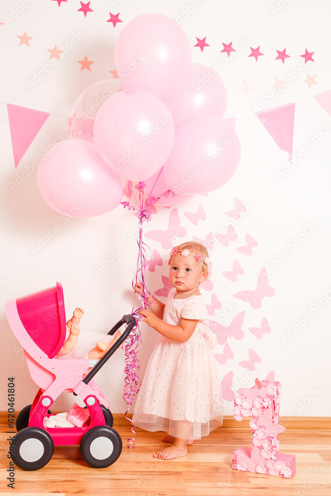 Happy blonde girl in a beautiful pink dress plays with doll's stroller at home. one-year photo shoot Happy blonde girl in a beautiful pink dress plays with doll's stroller at home. one-year photo shoot