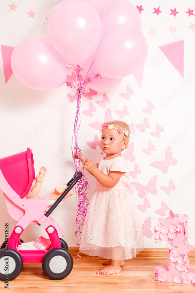 Happy blonde girl in a beautiful pink dress plays with doll's stroller at home. one-year photo shoot Happy blonde girl in a beautiful pink dress plays with doll's stroller at home. one-year photo shoot