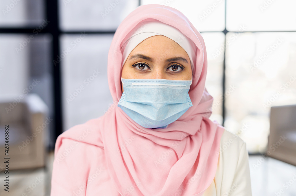Close up head shot portrait young serious cautious arabic ethnic woman employee worker wearing protective medical mask, looking at camera, keeping corporate quarantine rules, healthcare concept Close up head shot portrait young serious cautious arabic ethnic woman employee worker wearing protective medical mask, looking at camera, keeping corporate quarantine rules, healthcare concept