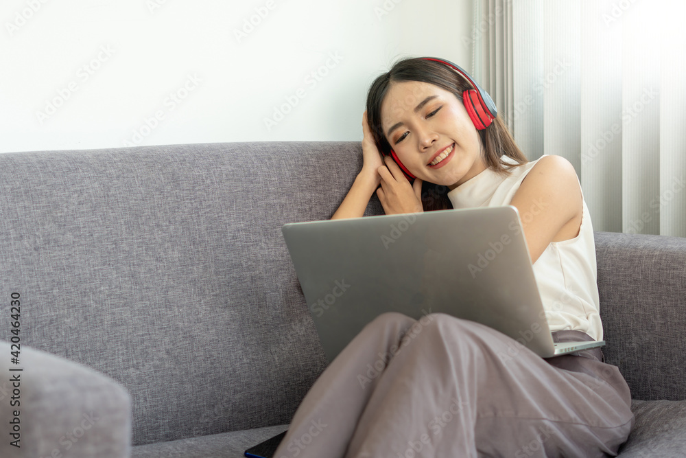 Naklejka premium A young Asian woman is happily relaxing listening to music through wireless headphones while at home on vacation.