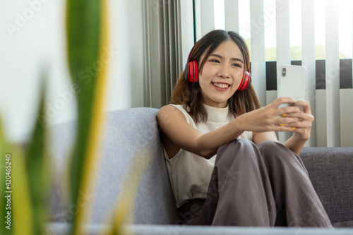 A young Asian woman wears headphones wirelessly, connects to a bluetooth system with a mobile phone. Chat with her friends in your free time while on vacation at home. #420465240