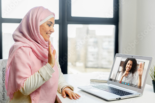 Young middle-eastern woman in hijab sits at the office desk, using app on PC for video call to coworker, colleague or friend, two young women in smart casual wear talking online via video connection