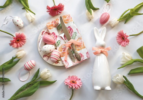 Easter table decoration with cutlery set  fork and knife on blooming napkin on roses plate  white tulips and pink daisy. Easter bunny and Easter eggs decor on white background  celebration concept