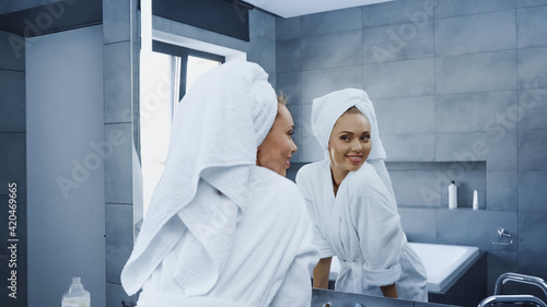 cheerful woman with towel on head looking at mirror in bathroom