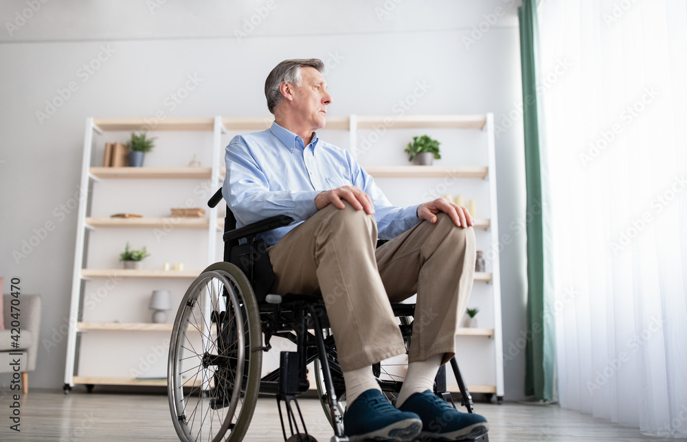 Low angle view of impaired male patient in wheelchair looking out window, feeling sad and desperate at home Low angle view of impaired male patient in wheelchair looking out window, feeling sad and desperate at home