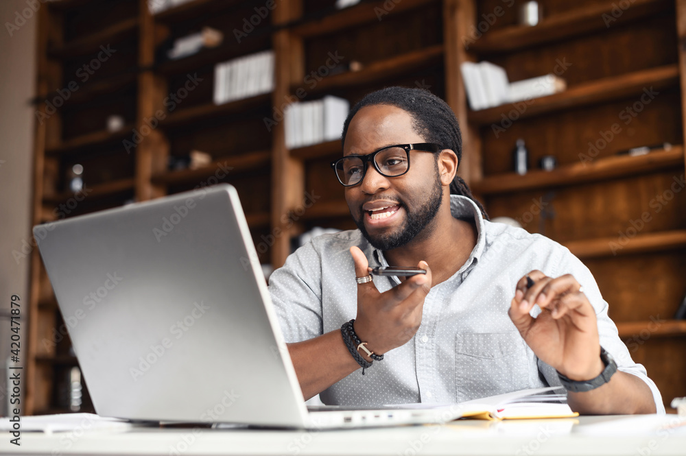 Busy African-American businessman in glasses with dreadlocks working in office, sitting at desk, holding smartphone, talking to customer on loudspeaker, looking at laptop screen, checking information