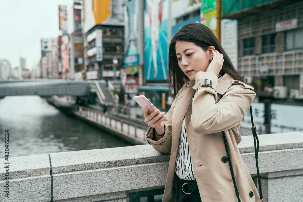 Young asian chinese woman travel in Osaka japan wearing jacket standing by river text on smartphone. dotonbori and shinsaibashi advertisement billboard area in background elegant girl using cellphone