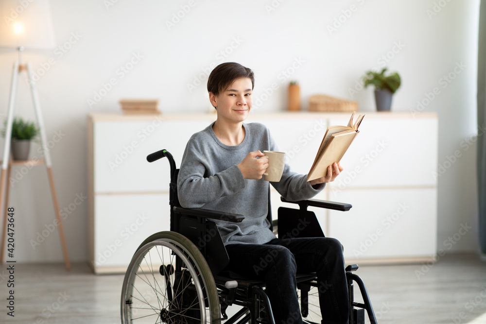 Positive impaired teenager in wheelchair drinking tea and reading interesting book at home