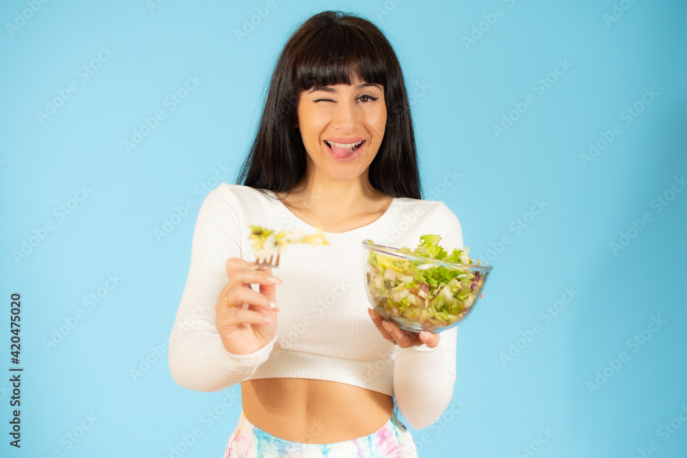 Portrait of a happy playful woman eating fresh salad from a bowl and winking isolated over blue background Portrait of a happy playful woman eating fresh salad from a bowl and winking isolated over blue background