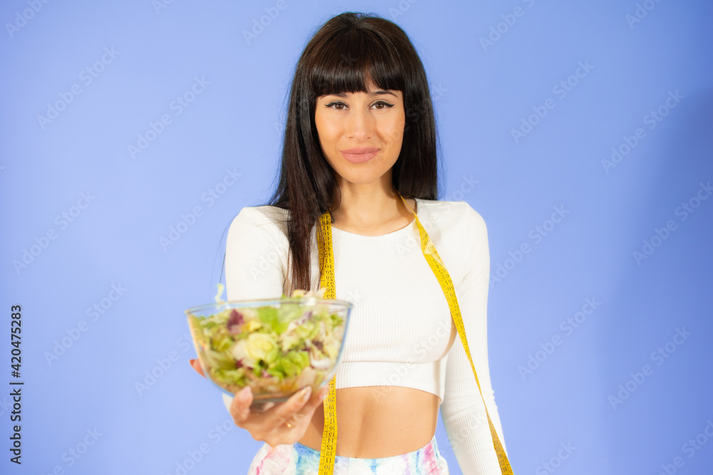 Healthy athletic woman in white sportswear with tape line on shoulders holding in hands bowl with vegetable salad Healthy athletic woman in white sportswear with tape line on shoulders holding in hands bowl with vegetable salad