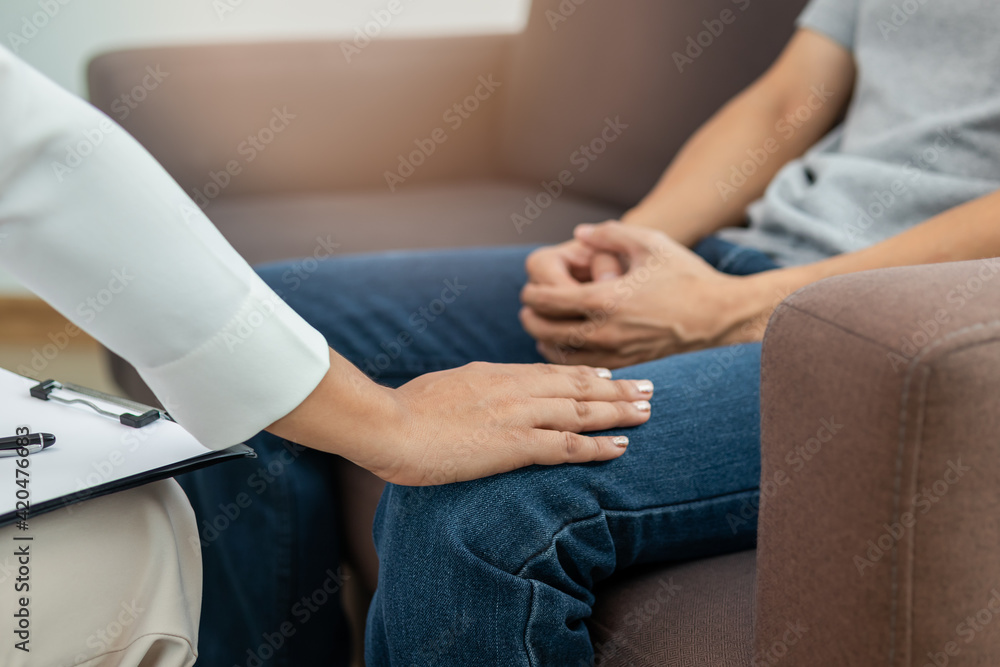 Young asian man, male suffers from a mental who needs to therapy with a psychologist while sitting on couch to consult, psychiatrist has encouragement the patient by touching to make his feel relaxed.