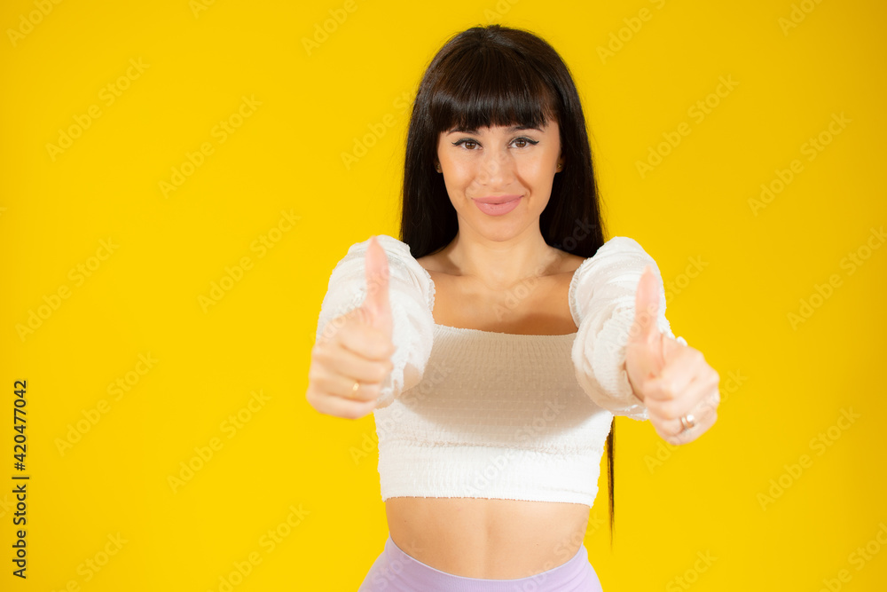 Attractive young woman giving a thumbs up gesture of approval and success over yellow background. Attractive young woman giving a thumbs up gesture of approval and success over yellow background.